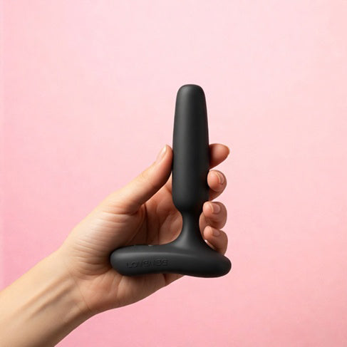 Hand holding Lovense Glint Touch-Responsive Vibrating Butt Plug Black against a pink background.